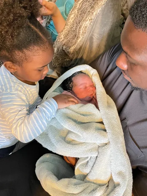 A parent and an older child looking down at a swaddled newborn