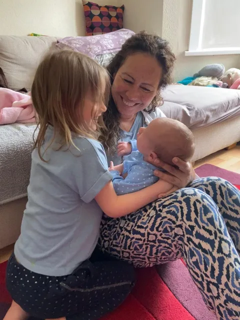 Regina smiling with an older child as they hold a newborn together
